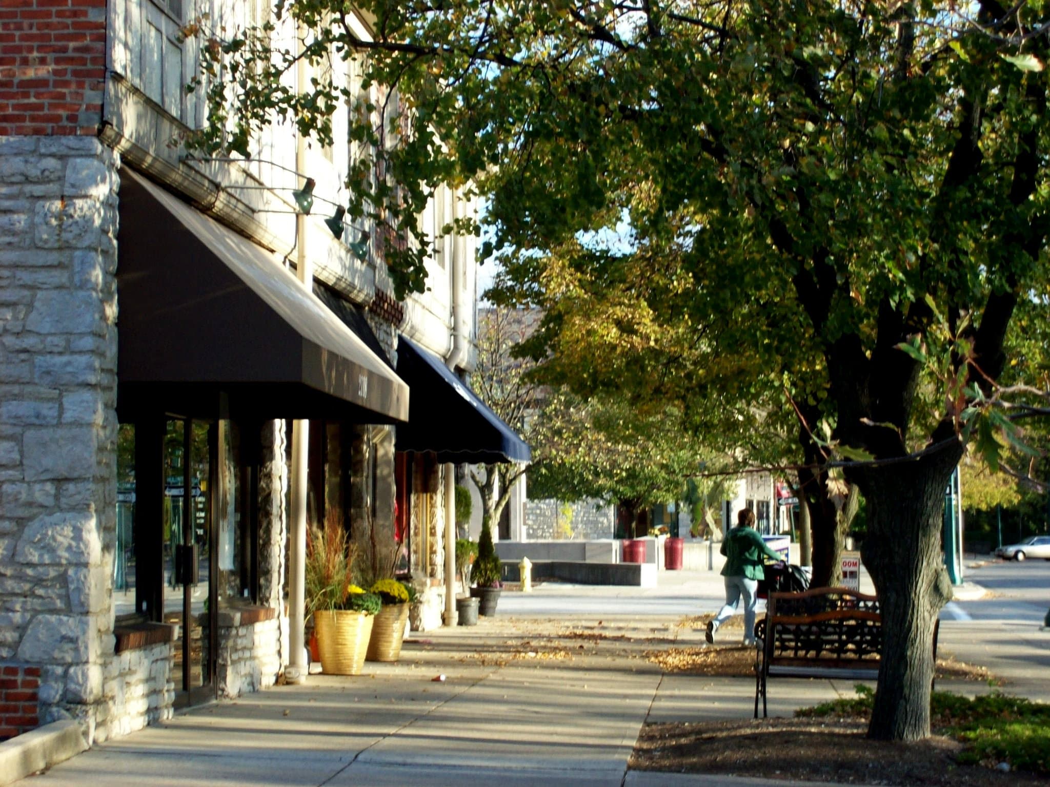 A tree-lined sidewalk with stone storefronts, awnings, and planters in Upper Arlington's historic Mallway shopping district